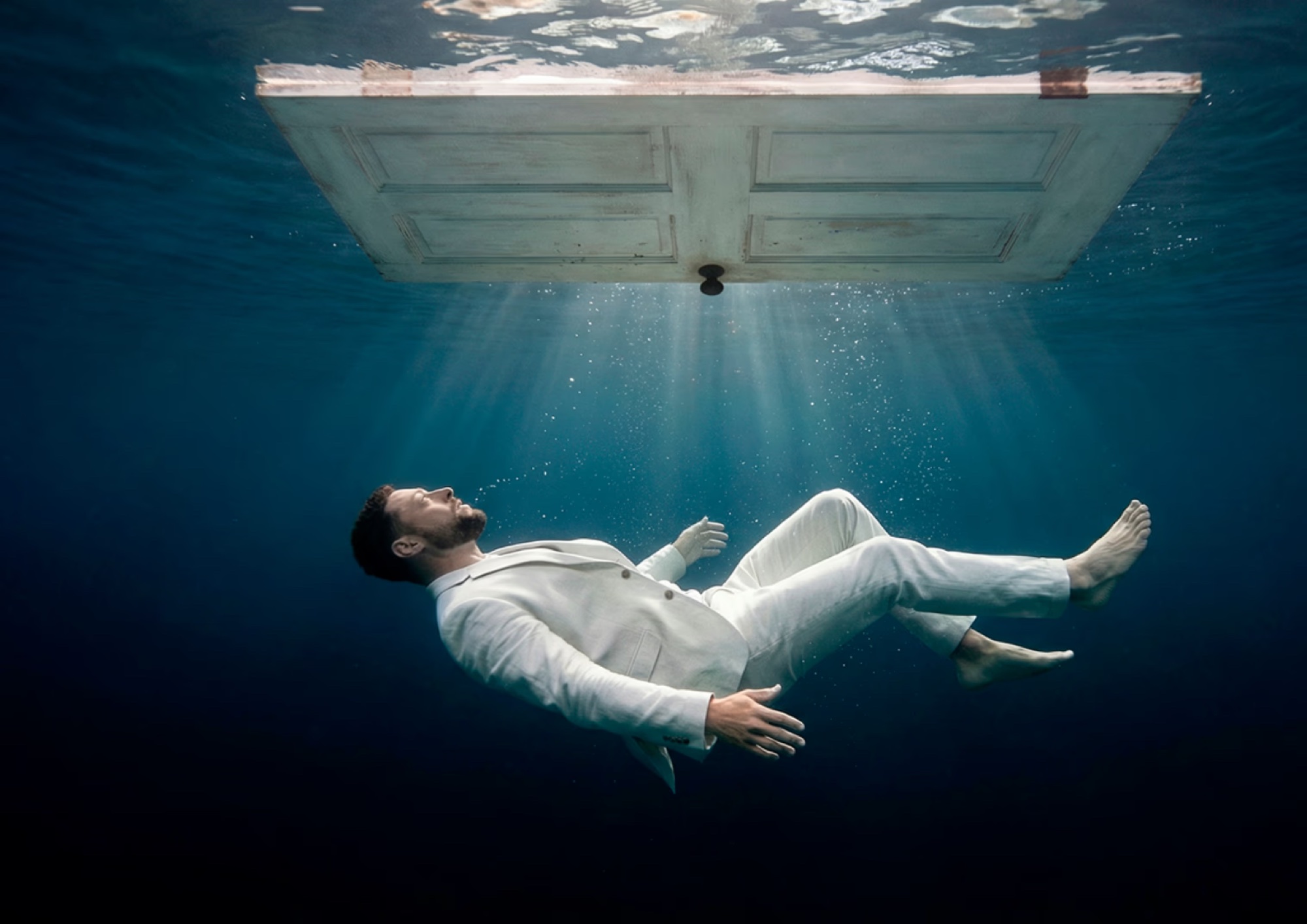 A man in a white suit suspended beneath a doorway of light, underwater